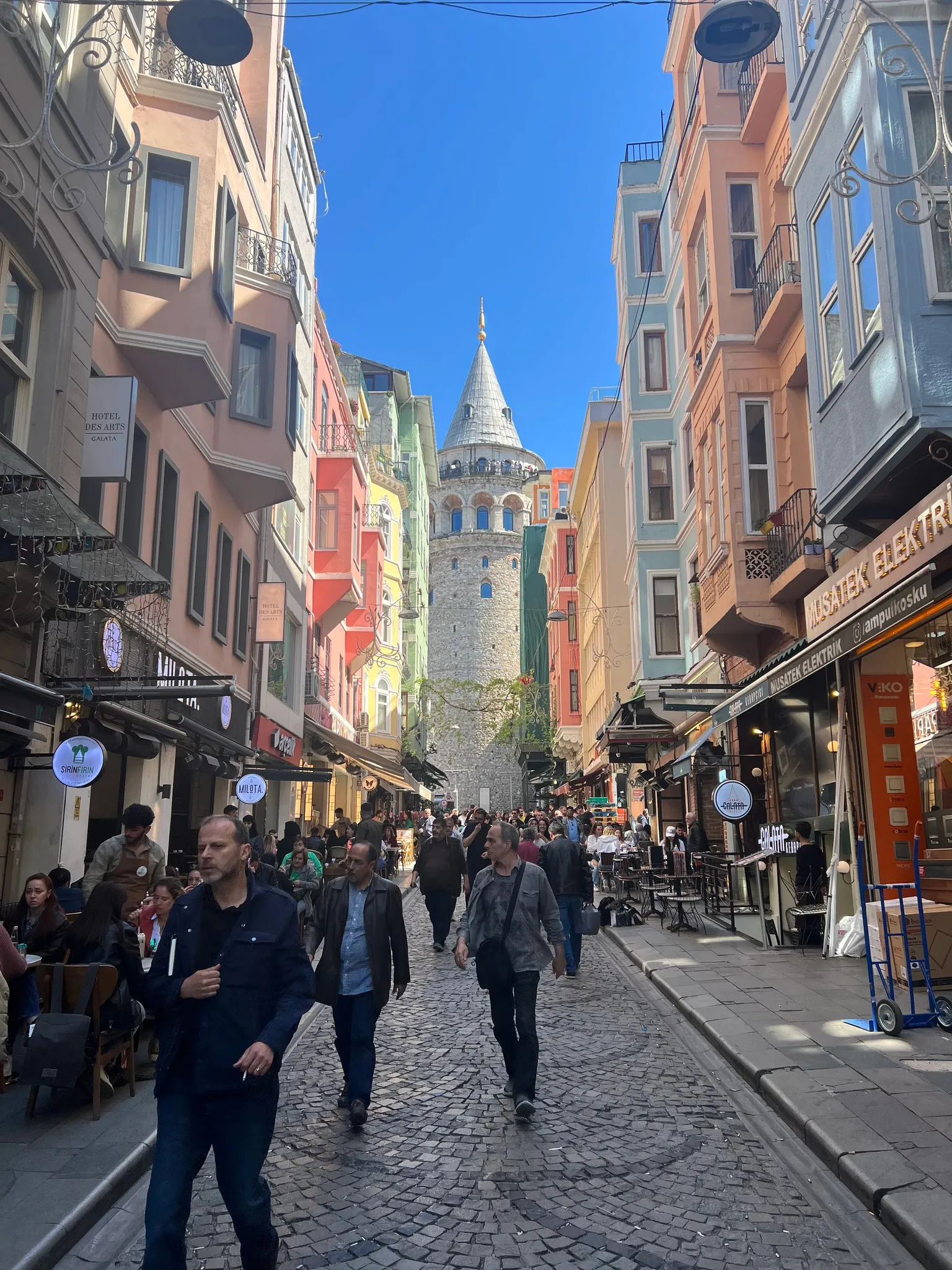 Vue d'une rue pavée animée à Istanbul, menant à la tour de Galata, avec des bâtiments colorés et des gens marchant.